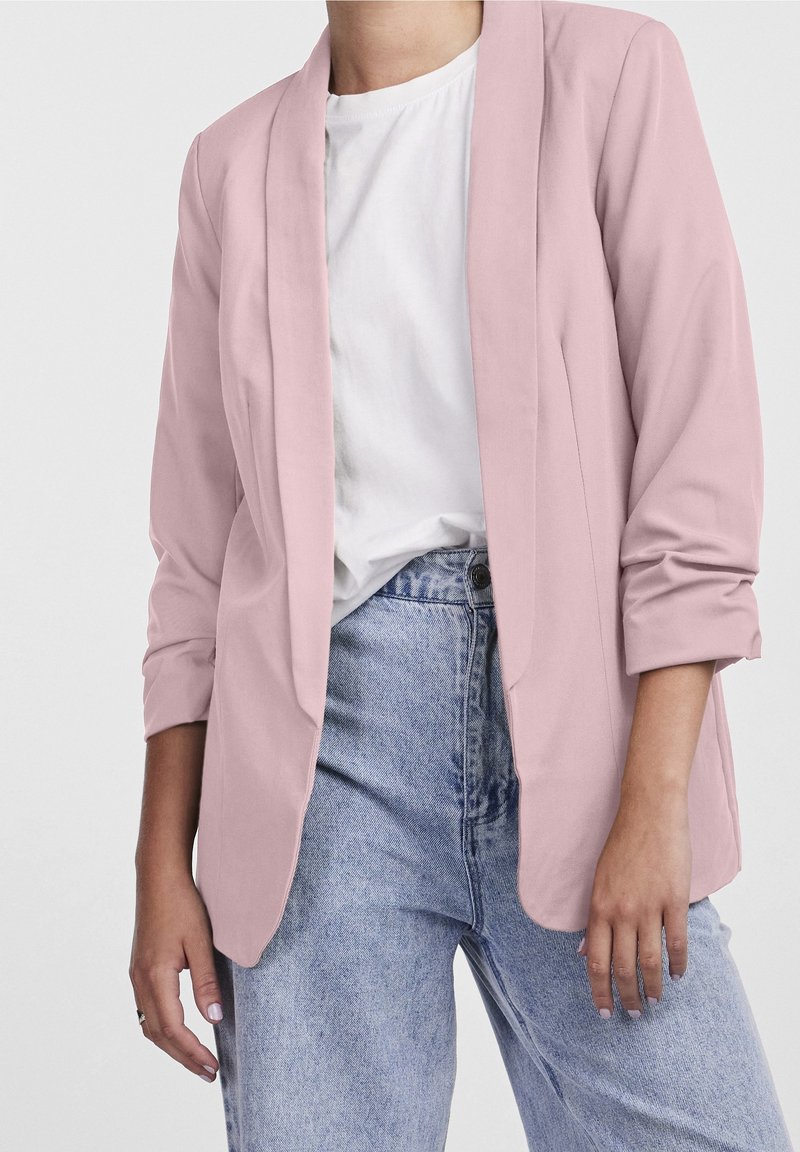 Femme portant un blazer rose pâle par-dessus une chemise blanche et un jean taille haute bleu clair, avec les manches remontées de façon décontractée.
