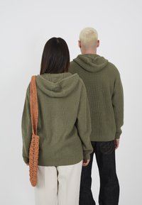 Olive green knit hoodies with ribbed texture, oversized fit. One model carries a woven orange bag. Black jeans feature star embroidery.