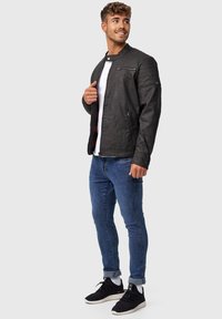 Schwarze Leder-Bomberjacke mit hohem Kragen, Reißverschlussfronttasche und gerippten Schulterakzenten. Kombiniert mit blauen Jeans und schwarzen Sneakers.