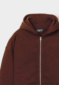 Brauner Reißverschluss-Hoodie mit weicher Textur, ausgestattet mit Kapuze, Fronttaschen und einem silbernen Reißverschluss, innen mit einem "BURBCS"-Schild versehen.