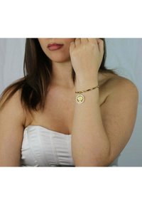 Bracciale tono oro con design attorcigliato, dotato di un ciondolo circolare inciso con un motivo ad albero, appoggiato su un polso nudo.