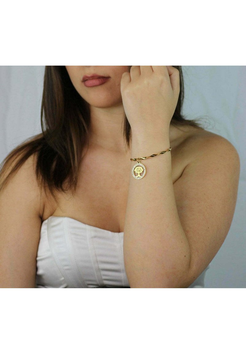 Bracciale tono oro con design attorcigliato, dotato di un ciondolo circolare inciso con un motivo ad albero, appoggiato su un polso nudo.