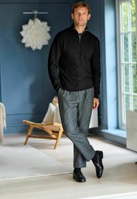 Czarny sweter robiony na drutach z teksturowanym wzorem w diamenty, szare spodnie z plisami i czarne buty eleganckie, w jasnym wnętrzu.