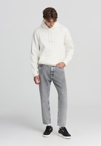 Hoodie crema a trama leggera con cordini, abbinato a jeans grigi chiari a gamba affusolata. Sneakers nere completano l'outfit. Design semplice e casual.
