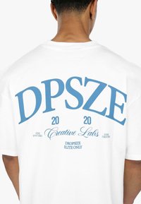 Hvid bomulds t-shirt med et stort blåt grafik på ryggen med teksten: "DPSZE 20 20 Creative Labs." Yderligere sætninger i mindre tekst nedenfor.
