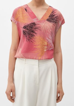 Vrouw draagt een roze top met V-hals met abstracte zwarte, oranje en witte veerachtige patronen, gecombineerd met witte hoge taillebroek.