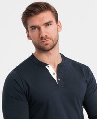 Man med kort brunt hår och skägg iförd en marinblå henley-tröja med tre träknappar på vit placket, mot en enkel ljus bakgrund.