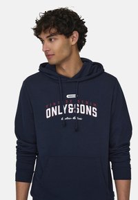 Sudadera de color azul marino hecha de tejido suave, con un bolsillo frontal, capucha con cordones y un atrevido gráfico de texto en blanco y rojo.