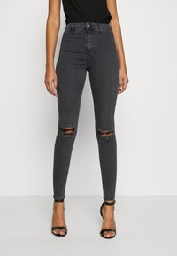 Grå högmidjade skinny jeans med rivna knän, gjorda av denimtyg. Har knappstängning och en figurnära design. Bärs med svarta klackar.