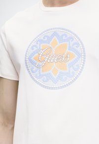 Άνδρας που φοράει λευκό T-shirt με κυκλικό μωσαϊκό λουλουδάτο σχέδιο σε παστέλ μπλε και ροδάκινο, με τη λέξη "Guess" σε καλλιγραφία στο κέντρο.