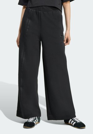 Partie inférieure du corps d'une personne portant un pantalon noir Adidas à jambes larges avec des rayures tonales et des baskets noires Adidas avec des rayures blanches.