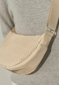 Beżowa teksturowana torebka na ramię z szerokim paskiem, noszona na szarej koszuli z długim rękawem.