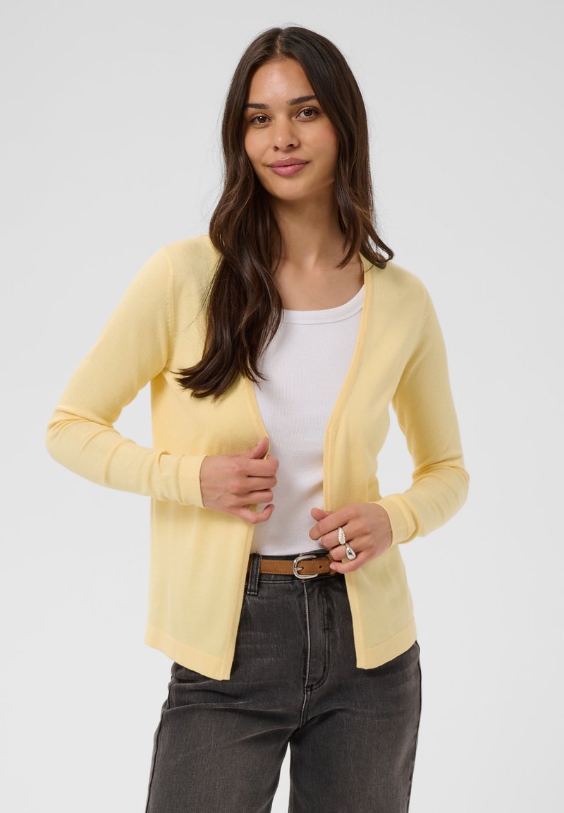 Jeune femme aux longs cheveux foncés portant un cardigan jaune clair sur un top blanc et un jean foncé, ajustant le cardigan avec ses deux mains.