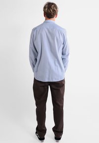 Chemise à manches longues bleu clair avec col, ourlet arrondi et texture subtile, associée à un pantalon marron foncé et des baskets noires.