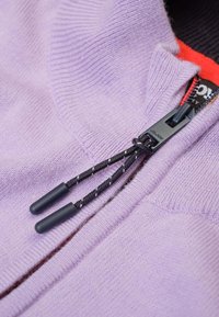 Lichtpaarse trui met een textuurbreiwerk, voorzien van een zwarte rits en koorden met rubberen uiteinden, en een binnenvoering in het rood.