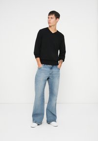 Czarny sweter z dekoltem w serek i długimi rękawami, w połączeniu z jasnoniebieskimi spodniami typu wide-leg. Model stoi z rękami w kieszeniach, nosząc białe buty.