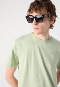 Black oversized sunglasses with angular frames and dark lenses. The person wears a light green cotton T-shirt with a round neckline.