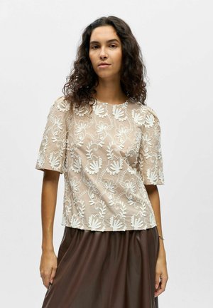 Femme aux cheveux bouclés foncés portant un haut beige à manches courtes avec broderies florales blanches et une jupe marron, debout devant un fond uni.