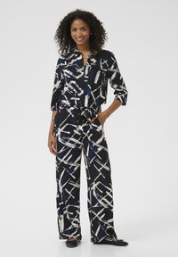 Black and white patterned jumpsuit with blue accents, featuring a collared shirt, pockets, a tie waist, and wide-leg trousers.