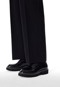 Black leather dress shoes with a glossy finish, featuring a decorative strap, white stitching, and a thick, textured sole.
