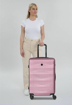 Mujer con camiseta blanca y pantalones beige de pie junto a una maleta rígida rosa con asa extendida sobre un fondo claro.