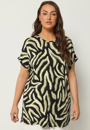 Vrouw met lang bruin haar die een zwart-olijfgroene tuniek met zebraprint en korte mouwen draagt, staand tegen een effen achtergrond.