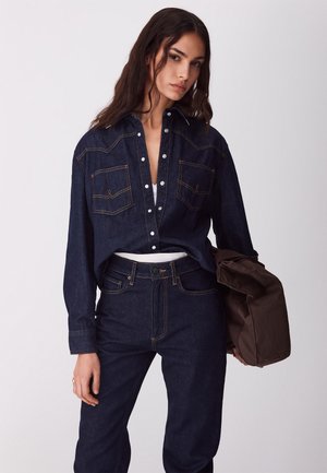 Kobieta ubrana w ciemnoniebieską koszulę dżinsową i jeansy, trzymająca brązową torbę w lewej ręce na jednolitym tle.