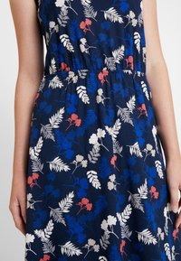 Robe florale en tissu bleu marine foncé, ornée d'imprimés botaniques blancs, rouges et bleus. Taille élastique, sans manches, texture douce.