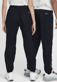 Črne lahke hlače za prosti čas z elastičnim pasom, stranskimi žepnimi zadrgami in zapenjanjem na gležnjih. Nike logo na hrbtni strani in nogi.