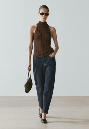 Giovane donna con top marrone senza maniche a coste, jeans scuri a vita alta, ballerine nere, occhiali da sole e una piccola borsa marrone in mano.