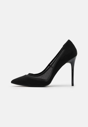 Sort højhælet pumpe med spids tå, mesh-sidesektioner og slank stiletthæl på en ensfarvet hvid baggrund.