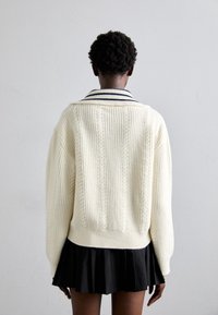 Grober, gestrickter cremefarbener Pullover mit Zopfmuster und einem marineblau gestreiften Kragen, kombiniert mit einem schwarzen Faltenrock. Weiche Textur und übergroße Passform.