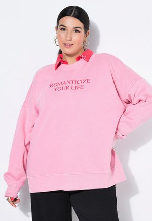 Kvinde med mørkt hår i en lav hestehale bærer en pink sweatshirt med rød tekst "ROMANTICIZE YOUR LIFE," rød skjorte med krave, sorte bukser og hoop-øreringe.