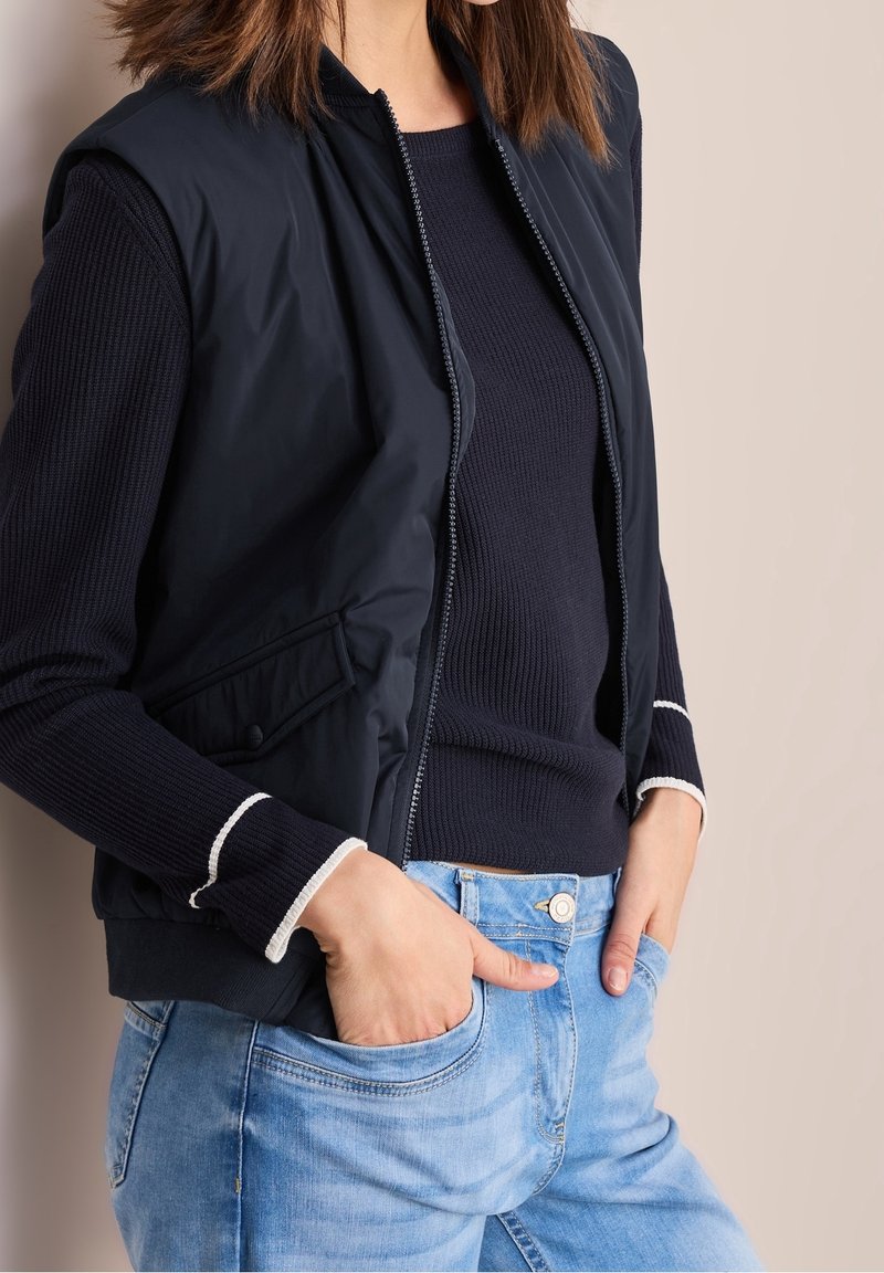 Femme portant un gilet sans manches bleu marine zippé sur un pull bleu marine avec une bordure blanche, et un jean bleu clair, les mains dans les poches sur un fond uni.