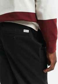 Schwarze Hosen aus glattem Stoff, mit einer Gesäßtasche mit einem Knopf und einem GANT-Label. Ein rot-weißer, gestreifter Langarm-Oberteil ist sichtbar.