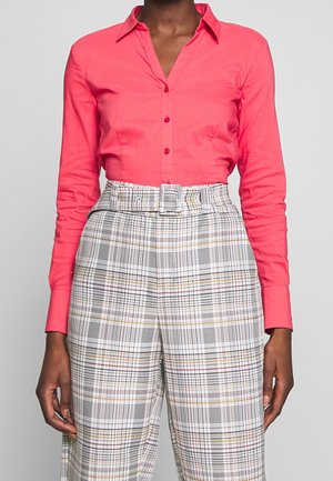 Femme portant un chemisier corail ajusté à boutons, rentrée dans un pantalon taille haute à ceinture, à carreaux blancs, noirs et tons atténués.