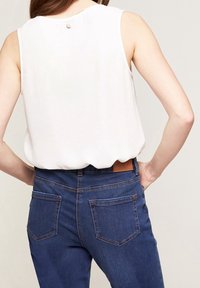 Weißes ärmelloses Oberteil mit einem Knopfdetails im Rücken, kombiniert mit dunkelblauen Jeans aus Denim mit sichtbaren Nähten und Patch-Akzenten.