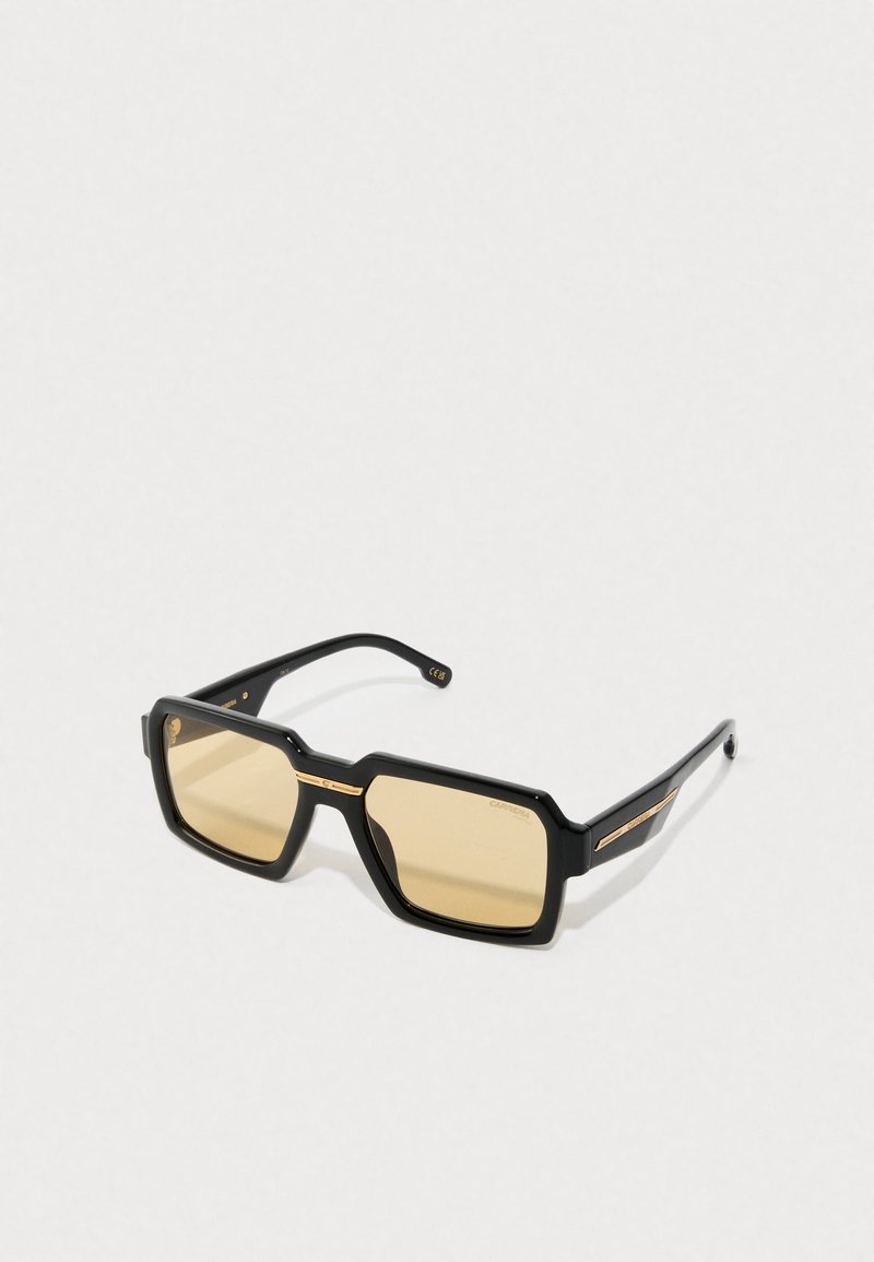 Óculos de sol pretos de plástico com armação quadrada e detalhes em dourado. Lentes amarelas. Braços texturizados com elementos decorativos.