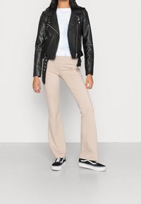 Zwarte leren motorjack met ritssluitingen, wit tie-dye shirt, beige uitlopende broek en zwarte sneakers met witte zolen.