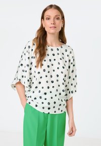 Locker sitzende, kurzärmelige Bluse in Weiß mit schwarzen runden Punkten, kombiniert mit leuchtend grünen Hosen. Glattes Material, lässiges Design.