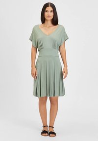Zielona sukienka typu jersey z dekoltem w serek, krótkimi rękawami i dopasowaną talią. Zwiewna spódnica do kolan o gładkiej fakturze i subtelnym układzie.