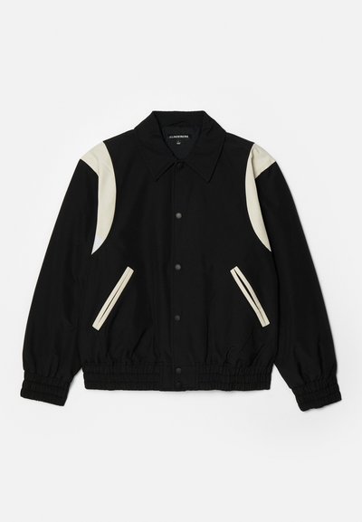 Black jacket with elastic cuffs, featuring cream-colored accents on shoulders and pockets. Snap-button closure, lightweight fabric, relaxed fit.