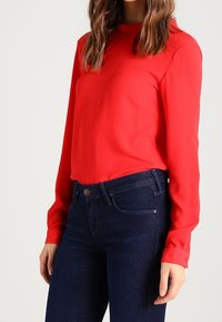 Röd långärmad blus med rund halsringning och slät textur, tillsammans med mörkblå tajta jeans med klassisk midja.