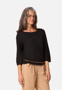 Zwarte top met driekwart mouwen van lichtgewicht stof, met brede halslijn, gecombineerd met beige broek, accentueert door een gouden kettingriem.