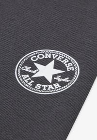 Melnas auduma virsma ar apļveida baltu logo, kurā ir zvaigzne un uzraksti "CONVERSE ALL STAR" un "Chuck Taylor". Teksturēta virsma ar šuvju detaļām.