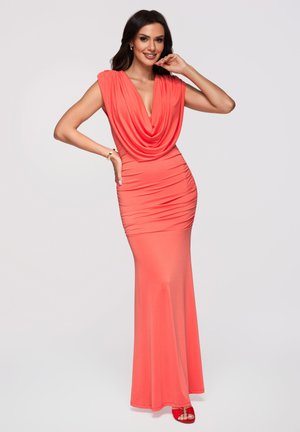 Femme portant une robe corail sans manches longue jusqu'au sol avec un décolleté drapé, posant avec une main sur la hanche et l'autre près du visage.