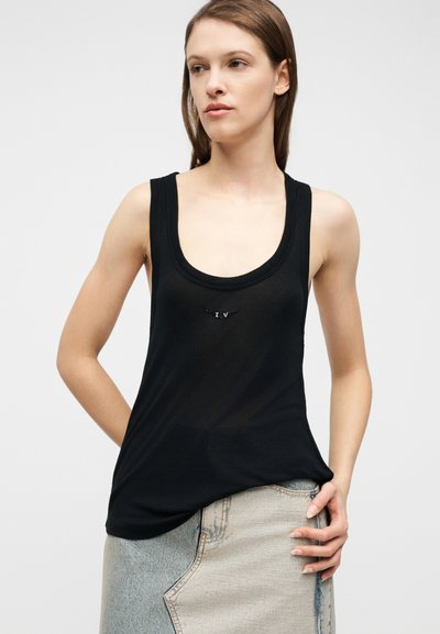 Νεαρή γυναίκα που φοράει μαύρο αμάνικο τοπ με μικρό λαμπερό λογότυπο και δίχρωμη τζιν φούστα, στέκεται μπροστά σε απλό φόντο.