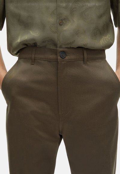 Pantalones marrones con una textura suave, que cuentan con un cierre de botón y bolsillos laterales. La camisa es de color verde oliva con un sutil patrón de paisley.