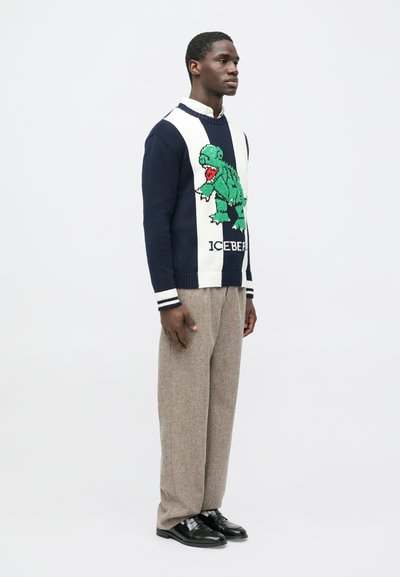 Άνδρας που στέκεται φορώντας μια μπλε ναυτικό και λευκή ζακέτα με έναν πράσινο δεινόσαυρο και το κείμενο "ICEBERG", μπεζ παντελόνια, λευκό πουκάμισο και μαύρα παπούτσια.
