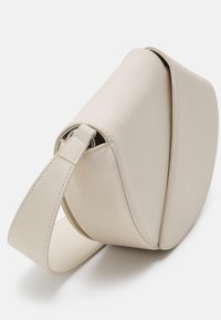 Borsa a tracolla in pelle crema con forma curva, tracolla piatta e design minimalista. Presenta una texture liscia senza hardware visibile.